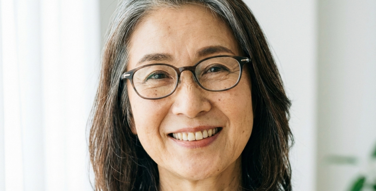 茶色いフレームのメガネをかけて白髪が混じったブラウンカラーのヘアスタイルをした60代の女性が微笑んでいる。